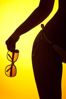 Silhouette of female body with bikini and sunglasses on yellow background, back lit