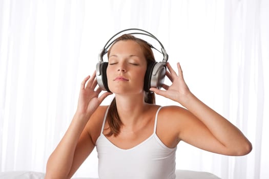 Young woman in white with headphones listening to music in bedroom