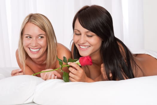 Two young smiling women naked in bed with red rose
