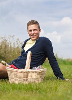 Young blond man sitting on the grass with picnic basket near him