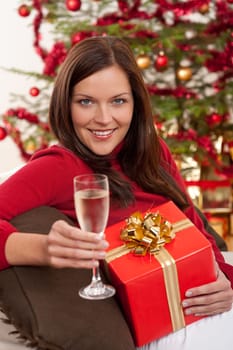 Smiling woman with Christmas present and glass of champagne in front of tree