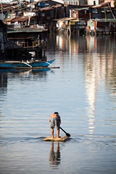 Man paddling down river on makeshift raft. Heavily polluted Paranaque River, Manila, Philippines. Squatter / Shanty housing lining the river banks.