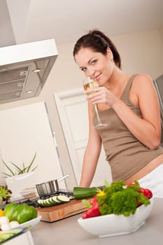 Smiling young woman cooking and holding glass of white wine in the kitchen