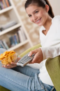 Students - Smiling female teenager watching television in living room with remote control and crisps