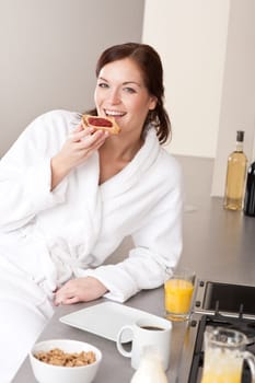 Young woman in bathrobe eating toast for breakfast in kitchen