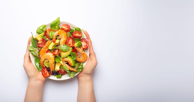Female hands holding salad with grilled shrimps, avocado, vegetables, green leaves on white plate isolated on white background top view. Clean eating, nutrition and dieting concept, copy space..