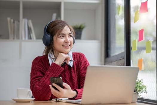 Asian female college student using laptop and phone with headphones while studying. Reading messages and greeting friends via video call.