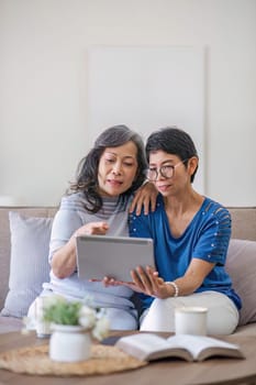 Two old female besties are watching a online media on a tablet while relaxing in the living room together..