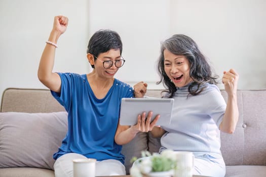 Two old female besties are watching a online media on a tablet while relaxing in the living room together..