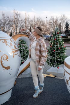 Woman Christmas Square. She stands near a large white cup, dressed in a light suit. With trees decorated with Christmas tinsel in the background.