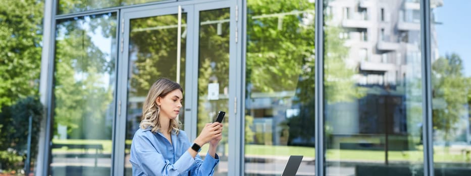 Portrait of businesswoman sitting outdoors and working. Young corporate woman looking at her smartphone, sitting outside with laptop and digital tablet.