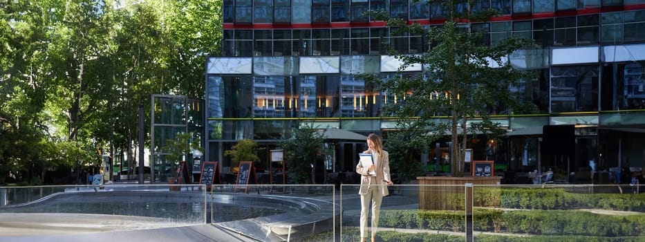 Corporate businesswoman in beige suit. Silhouette of young corporate woman with documents and laptop, posing outdoors in city center.