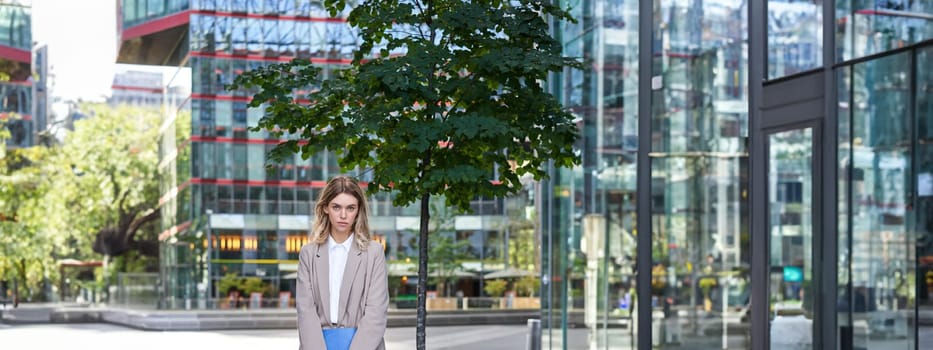 Portrait of young saleswoman in beige business suit, holding blue folder with work documents, standing outdoors on street of city center.