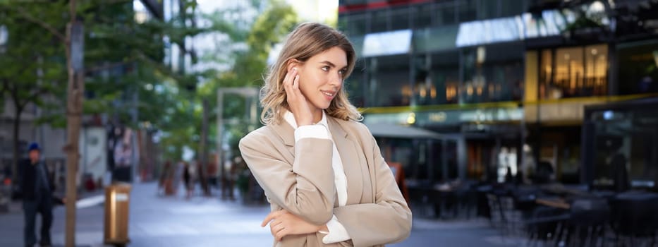 Close up portrait of blond businesswoman, confident corporate woman in headphones and beige suit, posing outdoors on street of city center.