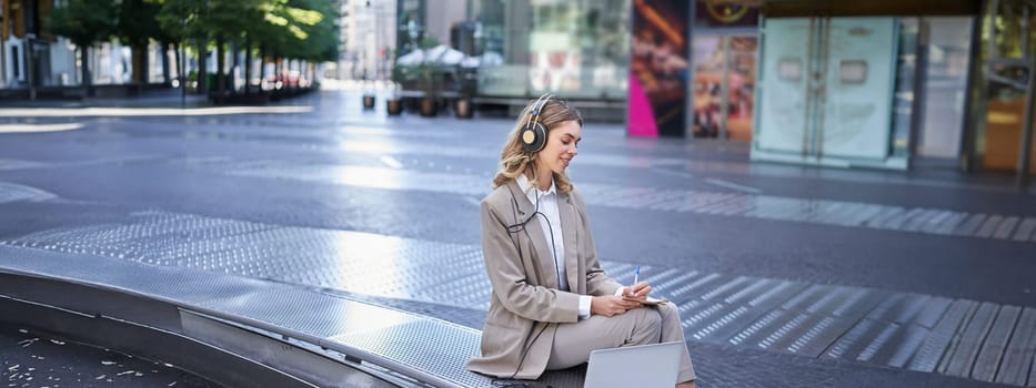 Woman sitting on a street with laptop and headphones plugged in, taking notes. Corporate worker attend online team meeting and writing down information, working outdoors.