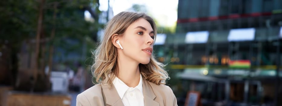 Close up portrait of blond businesswoman, confident corporate woman in headphones and beige suit, posing outdoors on street of city center.