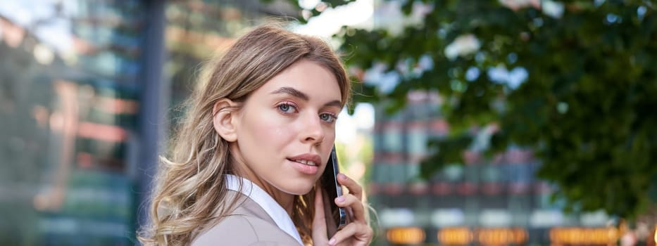 Close up shot of businesswoman talking on mobile phone. Corporate woman calling someone, looking around, standing outdoors.