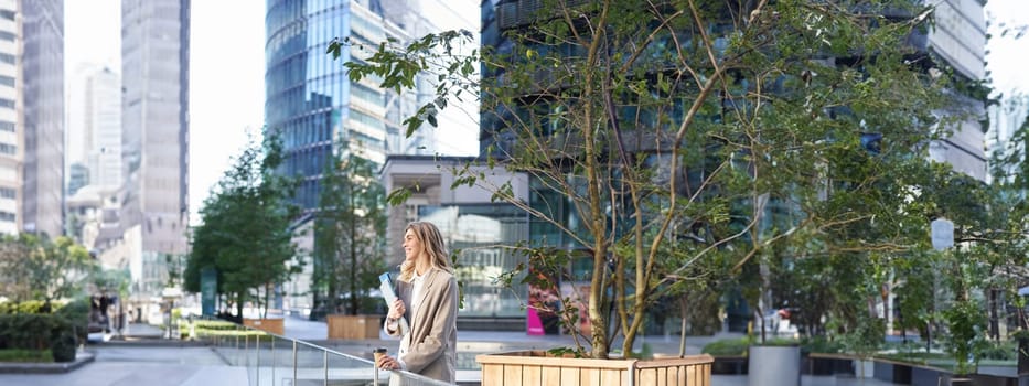 Vertical shot of smiling office girl in suit, drinks her coffee outdoors in city center, standing near office building, grabs takeaway.