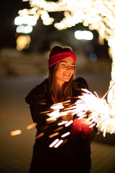 Woman holding sparkler night while celebrating Christmas outside. Dressed in a fur coat and a red headband. Blurred christmas decorations in the background. Selective focus.