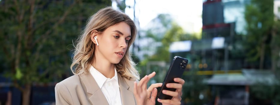 Successful businesswoman in beige suit, wireless headphones, looking at mobile phone, using smartphone app, standing outdoors on street.