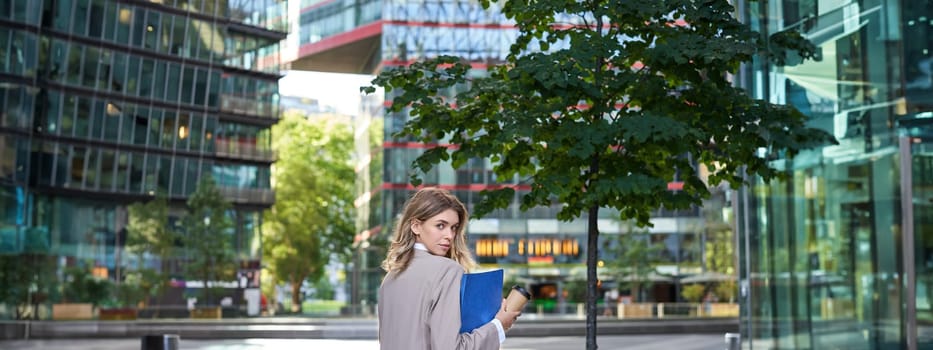 Silhouette of young business woman in beige suit, walking in city center, holding working documents and papers, standing outdoors near office buildings.