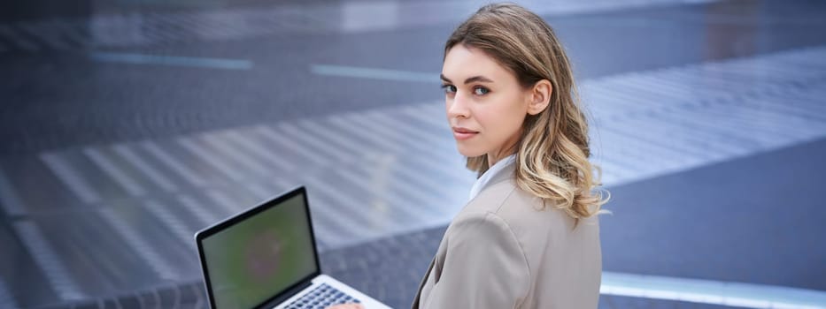 Young businesswoman using laptop while sitting outdoors in city centre, typing on keyboard. Girl preparing for interview, wearing suit.