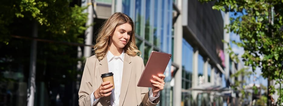 Portrait of young confident businesswoman on street, drinks her coffee and looks at tablet, works on her way to office. Digital nomad concept.