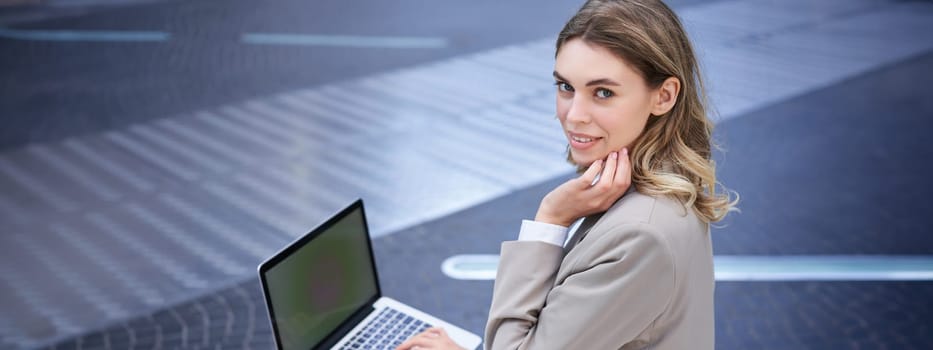 Young businesswoman using laptop while sitting outdoors in city centre, typing on keyboard. Girl preparing for interview, wearing suit.