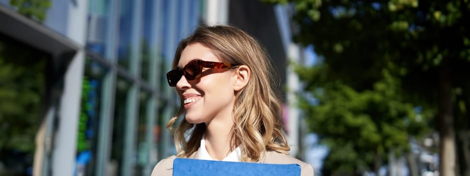 Close up portrait of smiling young woman in sunglasses, corporate suit, holding a folder with work documents, going to an interview, standing outdoors.