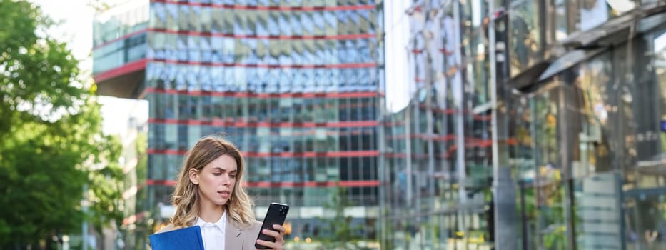 Vertical shot of corporate woman reading message on mobile phone, holding documents and drinking takeaway coffee, standing outdoors.