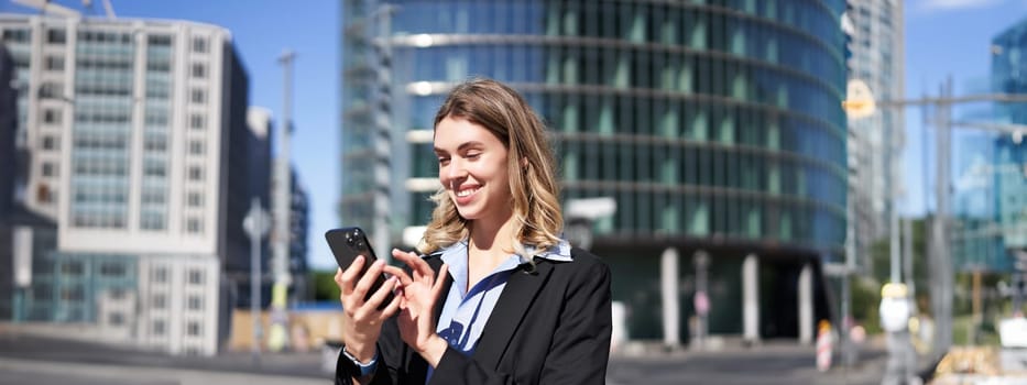 Portrait of confident corporate woman using mobile phone on city street. Businesswoman messaging on smartphone app while standing outdoors.