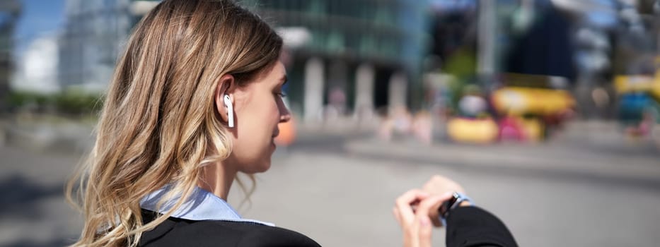 Portrait of busy corporate woman in wireless headphones, looking at time on her digital watch, checking messages, adjusting music, standing outdoors on street.