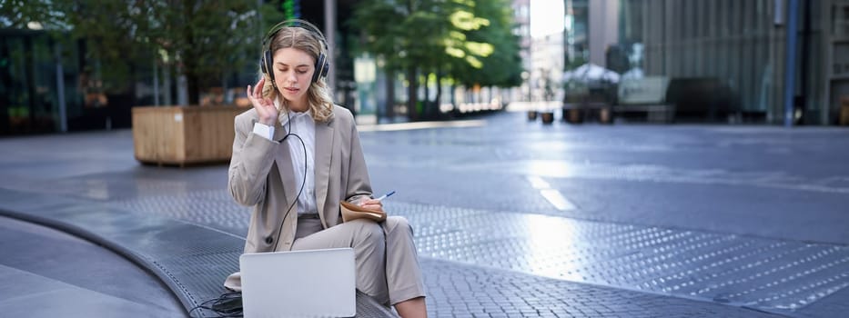 Woman sitting on a street with laptop and headphones plugged in, taking notes. Corporate worker attend online team meeting and writing down information, working outdoors.