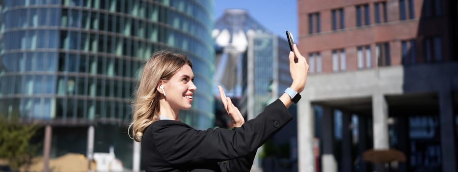 Portrait of businesswoman wave her hand at mobile phone camera, waves hand during video chat, stands in suit in city center outdoors.