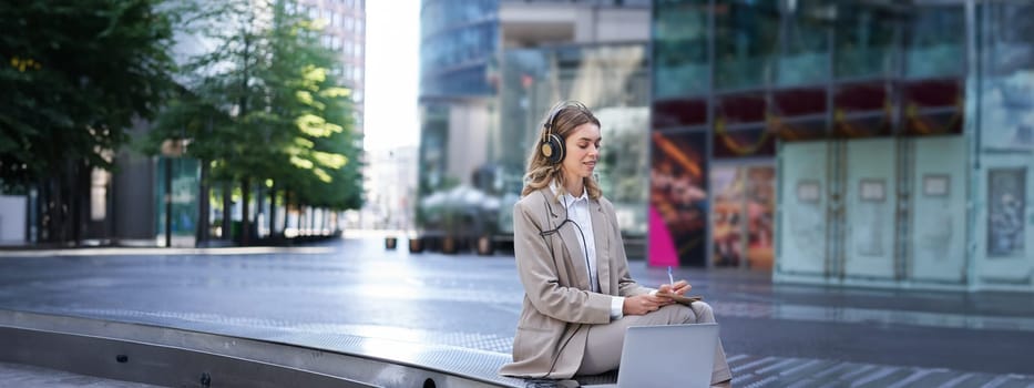 Woman sitting on a street with laptop and headphones plugged in, taking notes. Corporate worker attend online team meeting and writing down information, working outdoors.