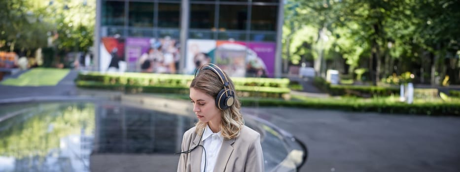 Portrait of businesswoman being on a meeting online outside office, listening in headphones, looking at laptop, working outdoors.