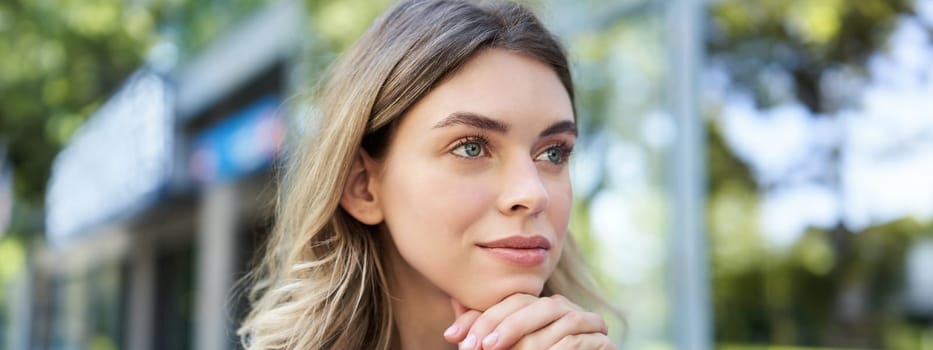 Close up portrait of young woman, looking hopeful and smiling, lean head on hands, thinking, sitting outdoors.