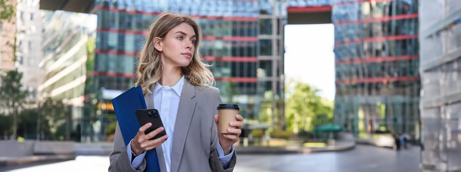 Businesswoman drinking coffee and holding documents with smartphone, going to work in office, standing outdoors in city center.