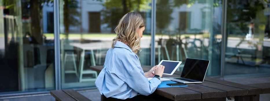 Portrait of businesswoman working on digital tablet, checking diagrams, sitting outdoors on fresh air near office building. Corporate woman prepare for work meeting.