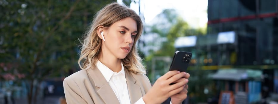 Successful businesswoman in beige suit, wireless headphones, looking at mobile phone, using smartphone app, standing outdoors on street.