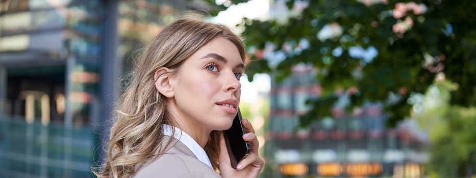 Close up shot of businesswoman talking on mobile phone. Corporate woman calling someone, looking around, standing outdoors.