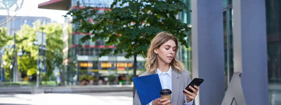 Businesswoman drinking coffee and holding documents with smartphone, going to work in office, standing outdoors in city center.