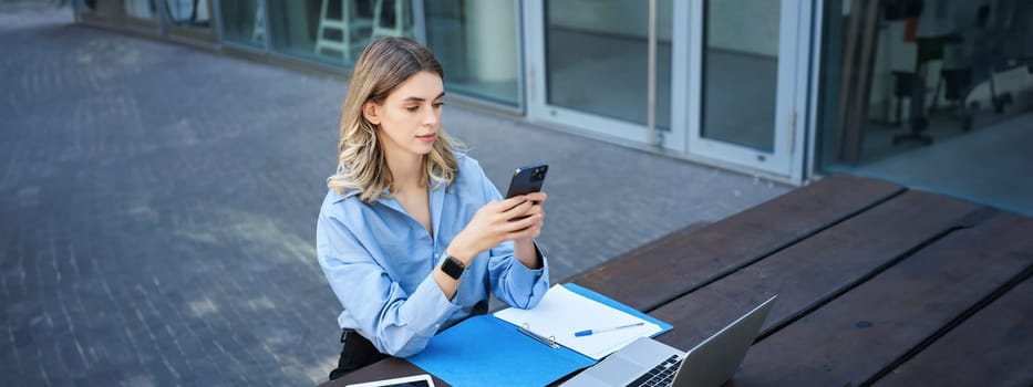 Portrait of businesswoman sitting outdoors and working. Young corporate woman looking at her smartphone, sitting outside with laptop and digital tablet.