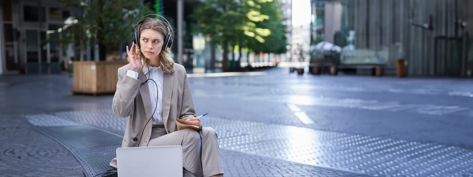 Woman sitting on a street with laptop and headphones plugged in, taking notes. Corporate worker attend online team meeting and writing down information, working outdoors.