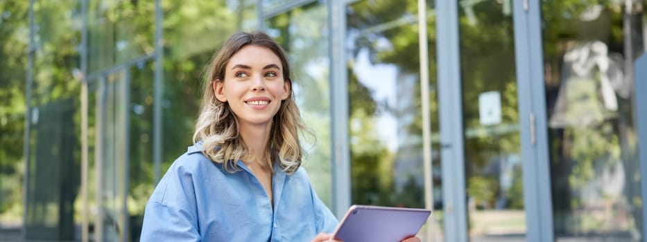 Young digital nomad, woman with tablet, working outdoors on fresh air, smiling and looking away at passerby.