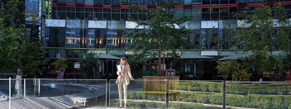 Corporate businesswoman in beige suit. Silhouette of young corporate woman with documents and laptop, posing outdoors in city center.