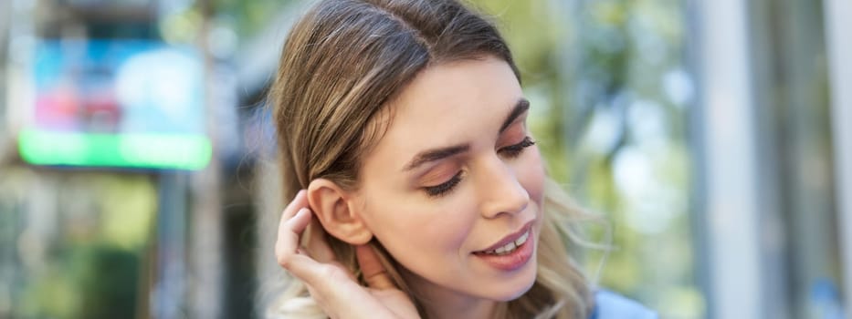 Close up portrait of young woman, tuck hair behind ear, looking flirty and smiling, sitting in blue shirt outdoors on street.