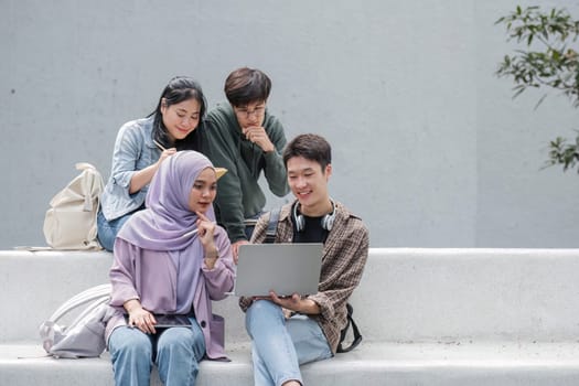 Group of happy young Asian college students sitting on a bench looking at a laptop screen, discussing and brainstorming on their school project together.