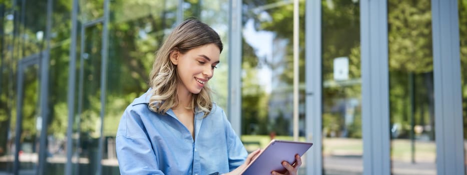 Young digital nomad, woman with tablet, working outdoors on fresh air, smiling and looking away at passerby.