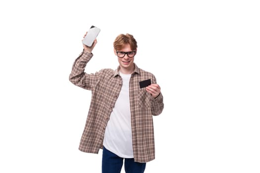 a young european man with red short hair and glasses is dressed in a plaid shirt holding a phone and a credit card.